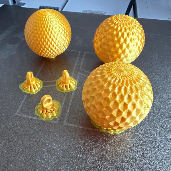 Tres boles de Nadal impreses en 3D en filament daurat, amb patrons calats diferents, i tres penjadors sobre els suports d'impressió